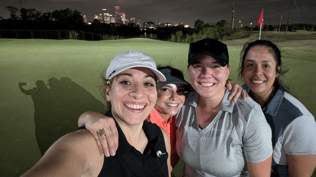 Four female golfers taking a selfie during a round of night golf in Houston Texas. Houston City Skyline in the background.